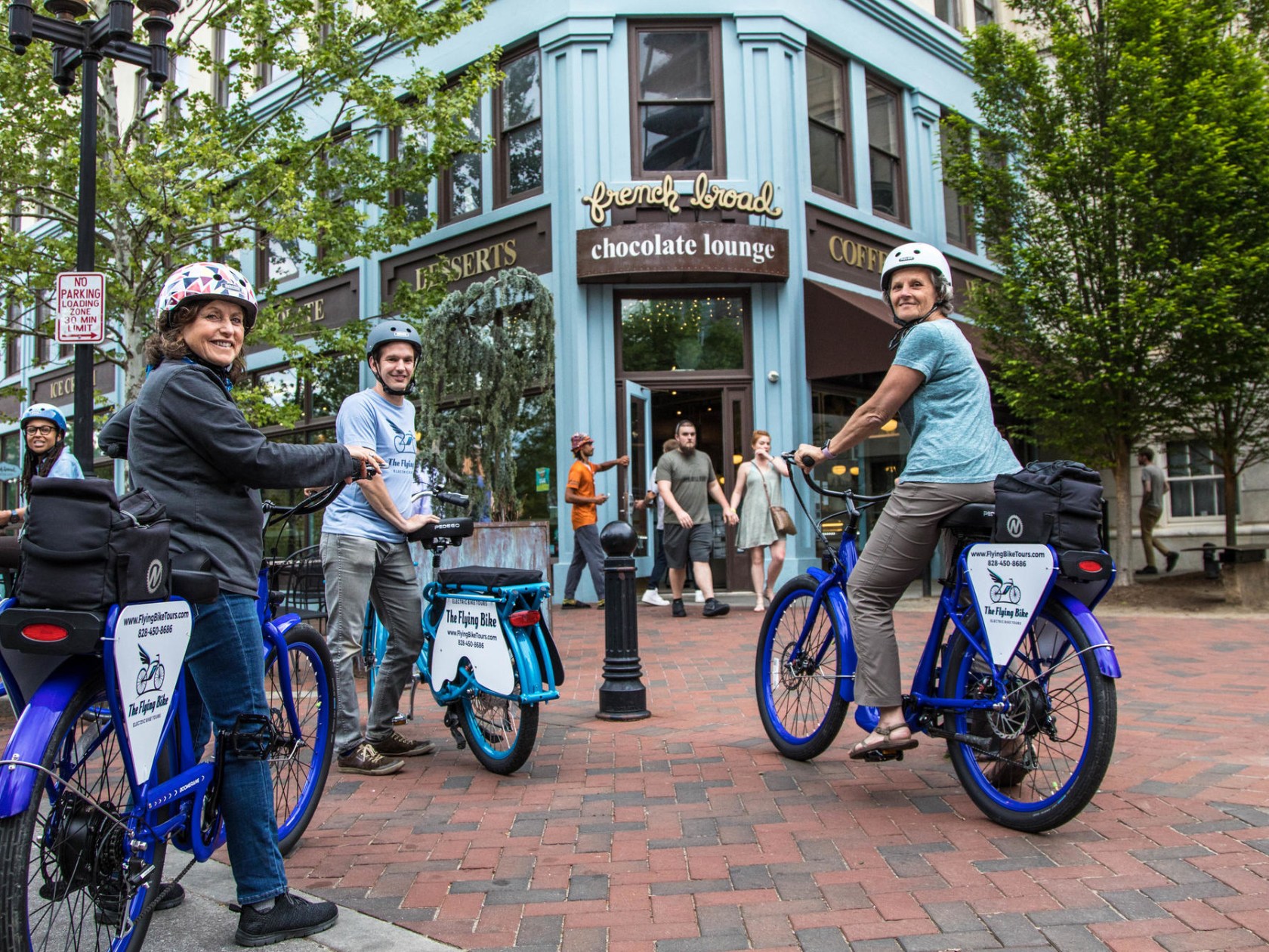 Group of people smiling with their electric bikes during a food tour of Asheville, NC. They are in front of a restaurant called the Chocolate Lounge on a brick paved sidewalk.