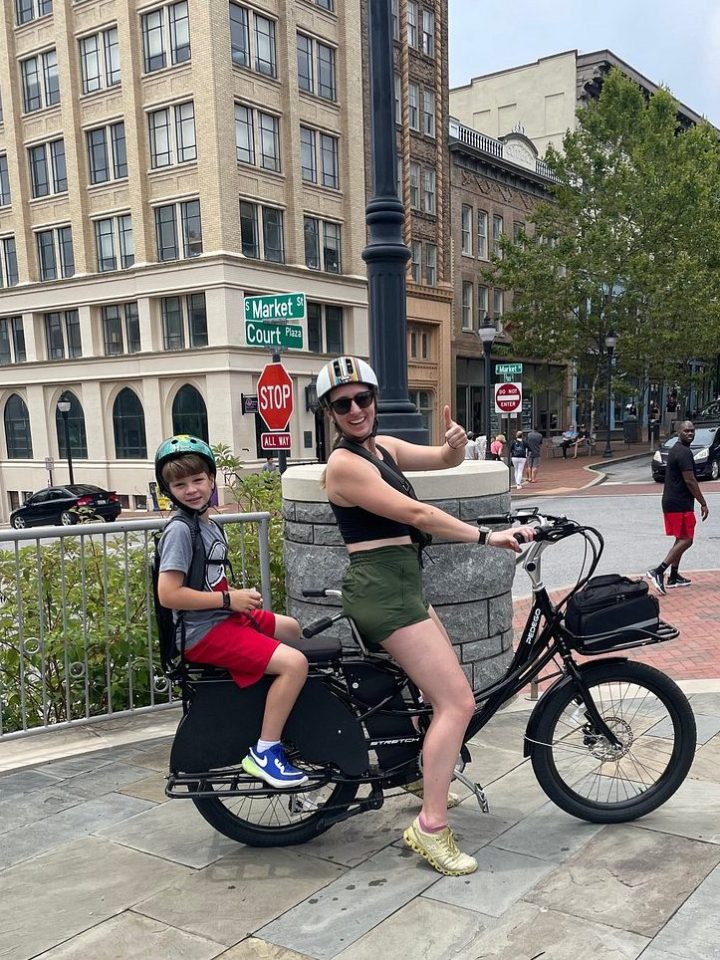 Woman and child on a cargo bike near a city street, both wearing helmets.