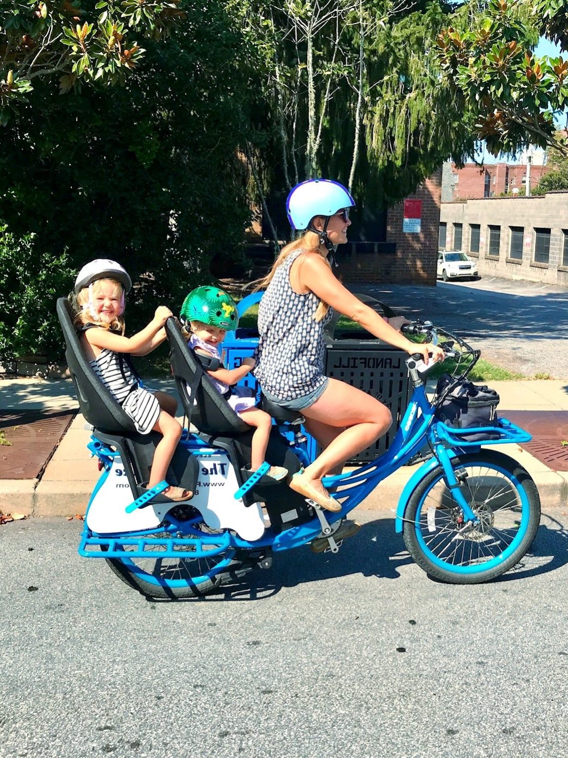 Woman biking with two children in seats on a sunny street.